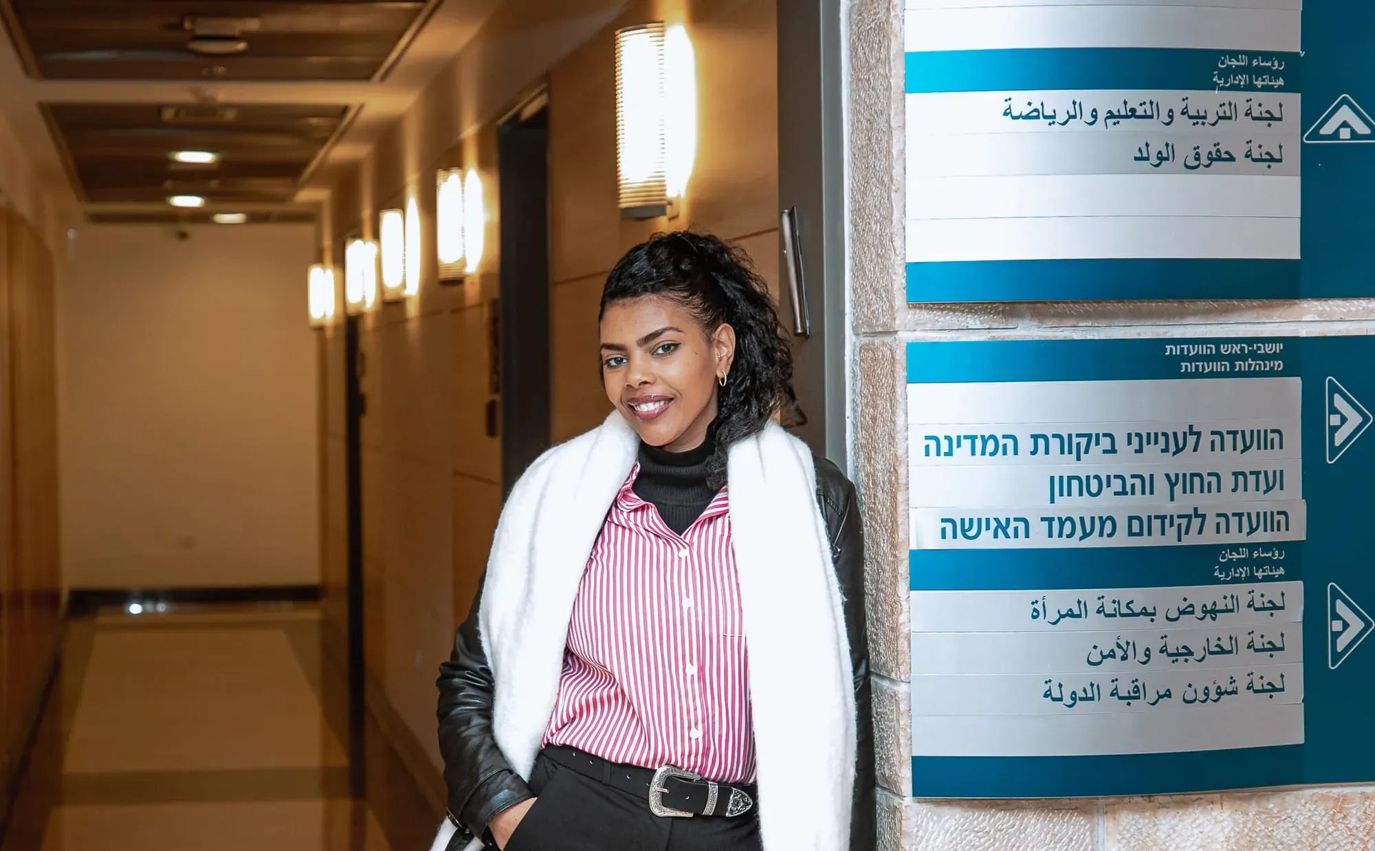 אשה במעיל לבן ומשקפיים עומדת במסדרון ליד שלטי ועדות