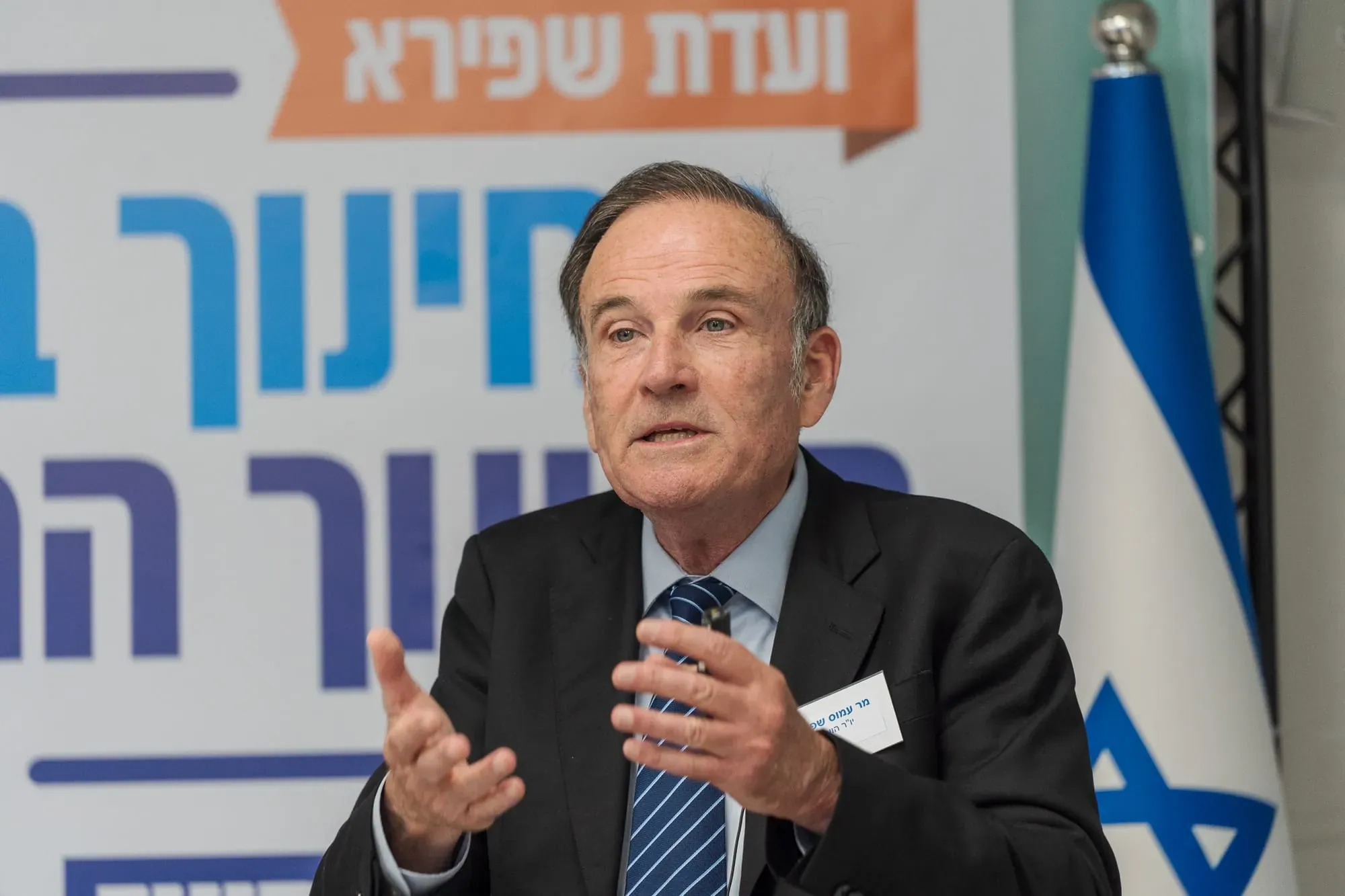 גבר מבוגר בחליפה מדבר בכנס עם שלט ועדה ודגל ישראל