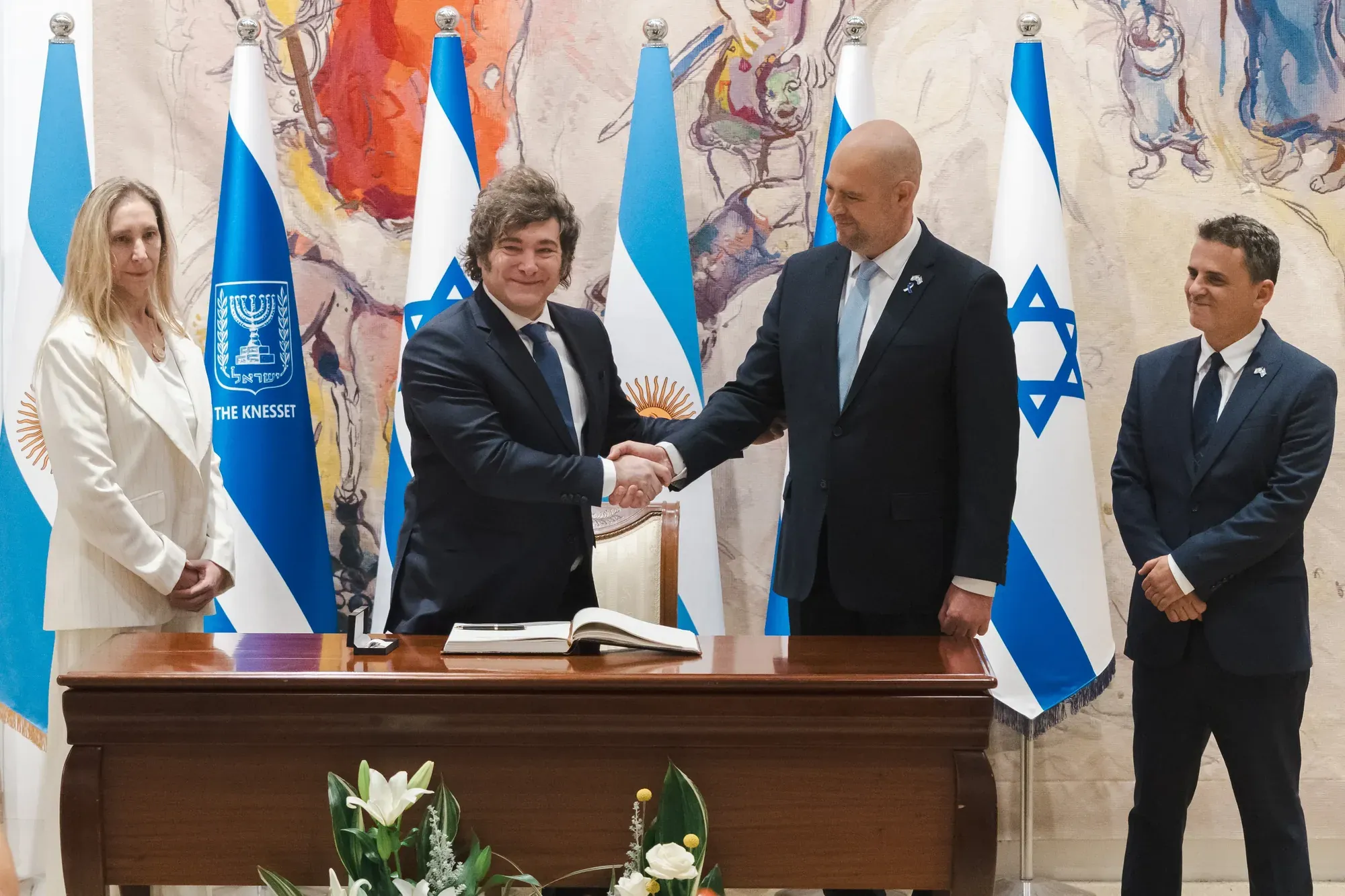 לחיצת יד וחתימה בספר אורחים מול דגלי ישראל וארגנטינה