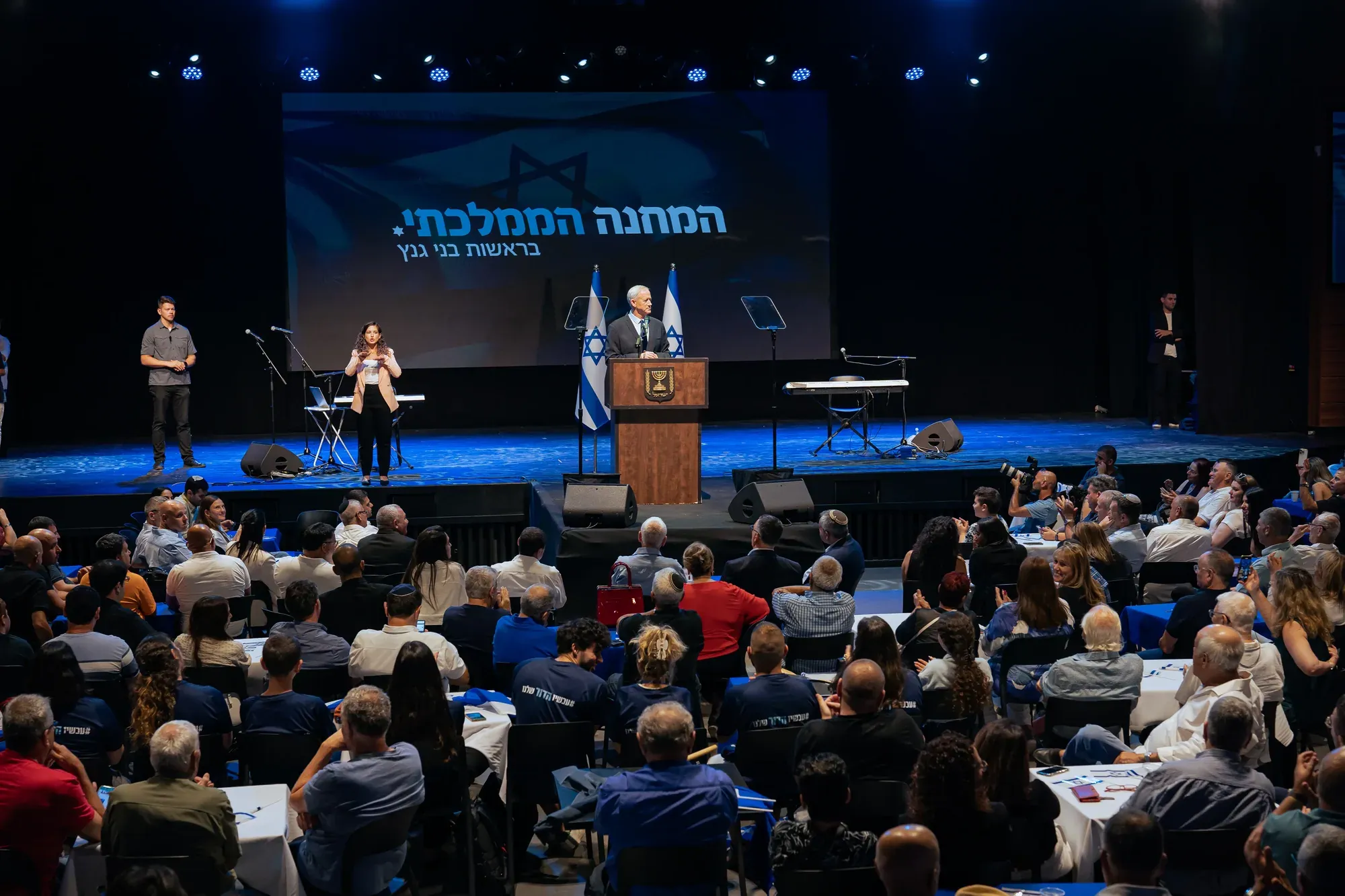 קהל בכנס פוליטי באולם, במה עם דגלי ישראל
