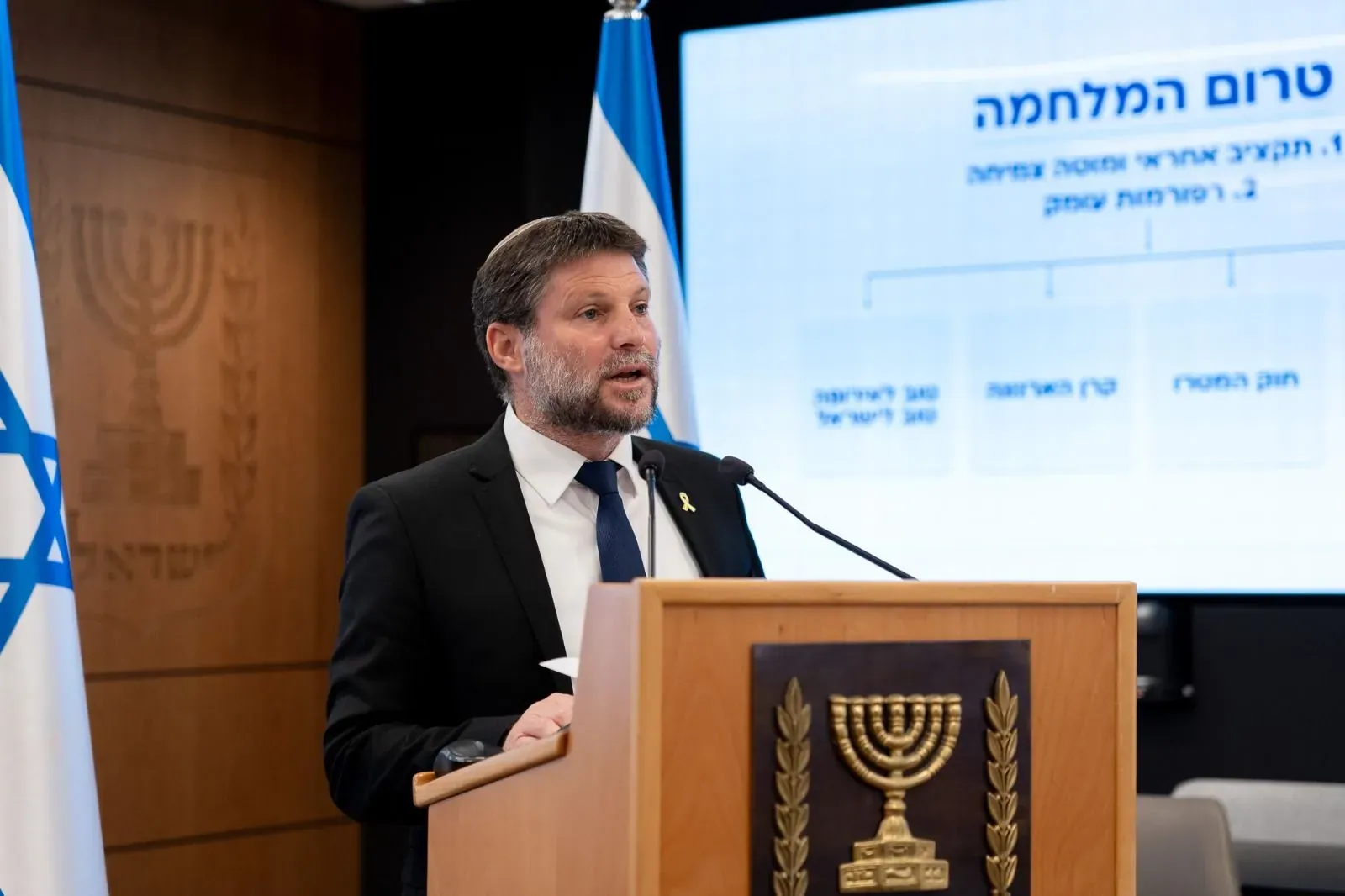 צילום אירוע: בצלאל סמוטריץ' נואם מול מצגת, ליד דוכן עם סמל המדינה
