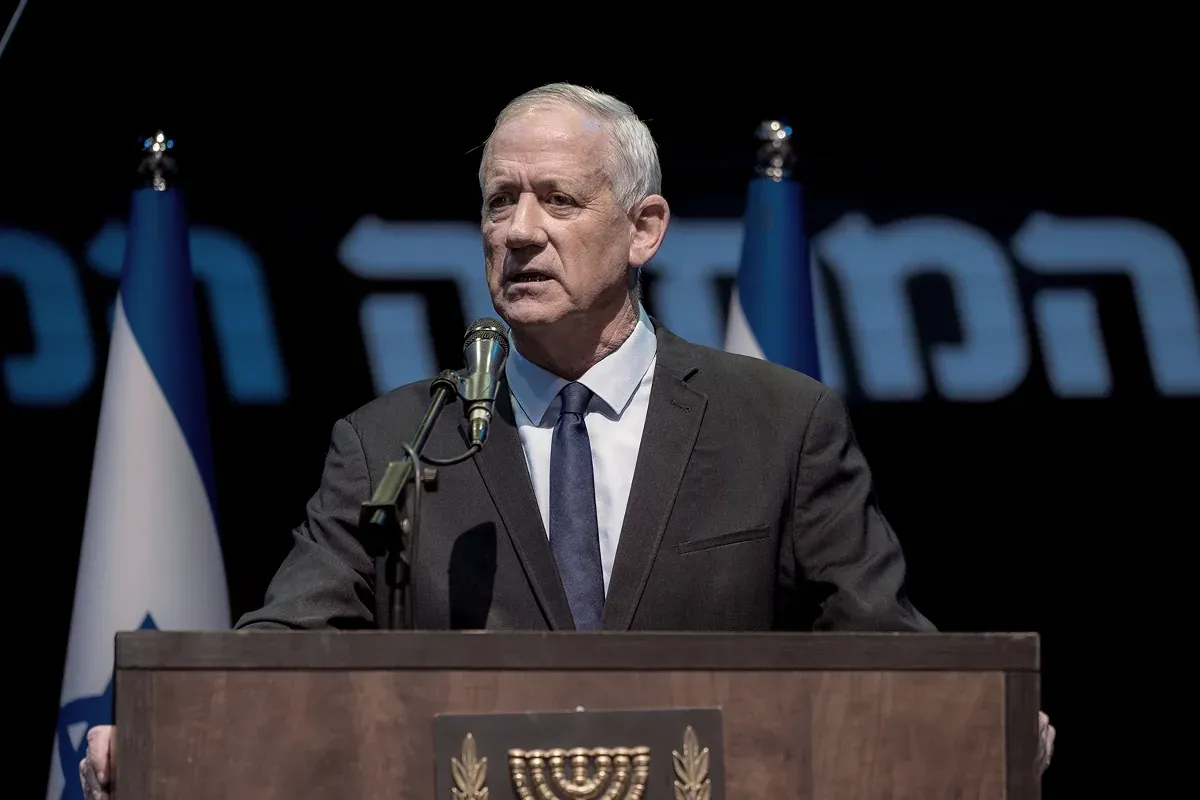 נואם בחליפה ליד דוכן עם סמל המדינה על רקע שלט בעברית