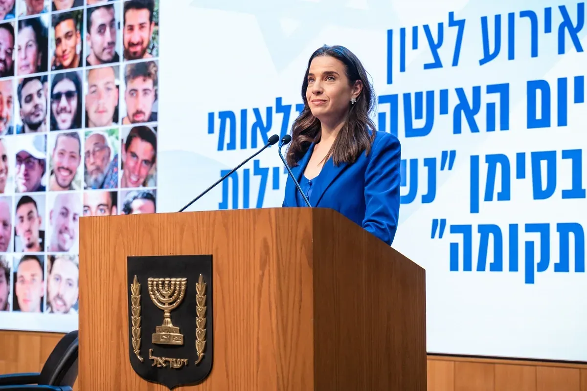 אשה בחליפה כחולה נואמת מאחורי דוכן באירוע לציון יום האשה הבינלאומי