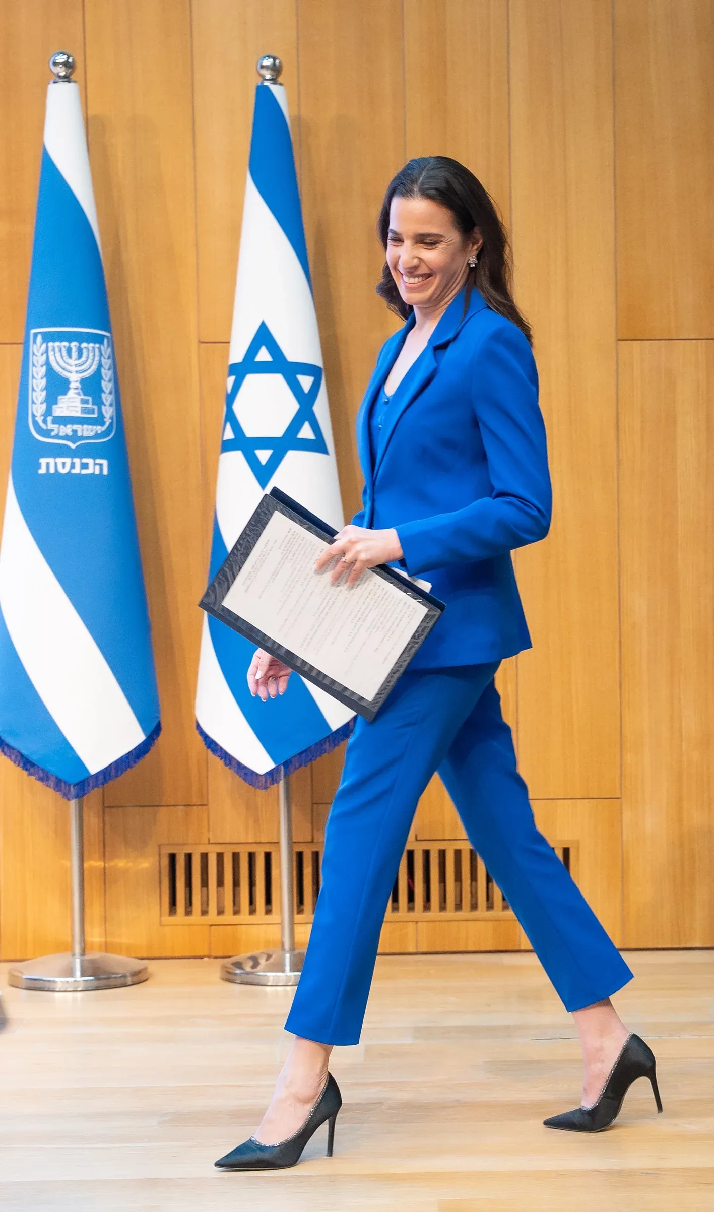 אשה בחליפה כחולה מחזיקה מסמך ופוסעת ליד דגלי ישראל
