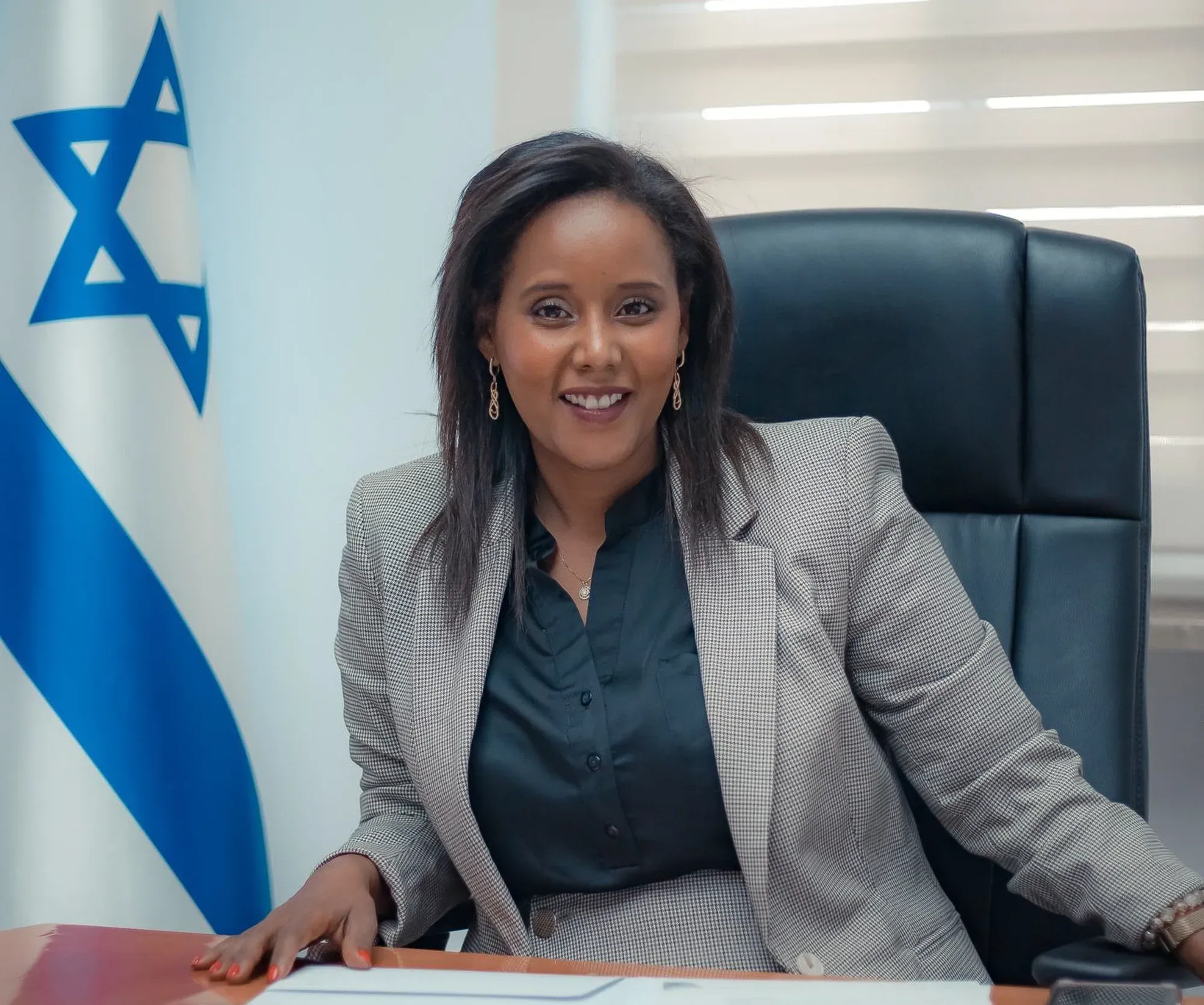 צילום תדמית: פנינה תמנו שטה יושבת במשרד בז'קט אפור לצד דגל ישראל