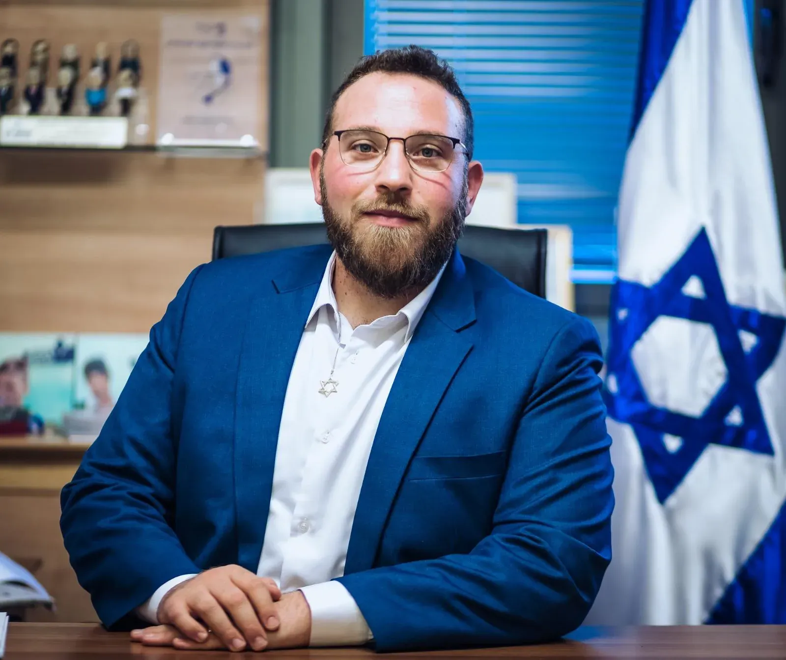 פורטרט עסקי של גבר עם משקפיים וזקן בחליפה כחולה ליד שולחן ודגל ישראל