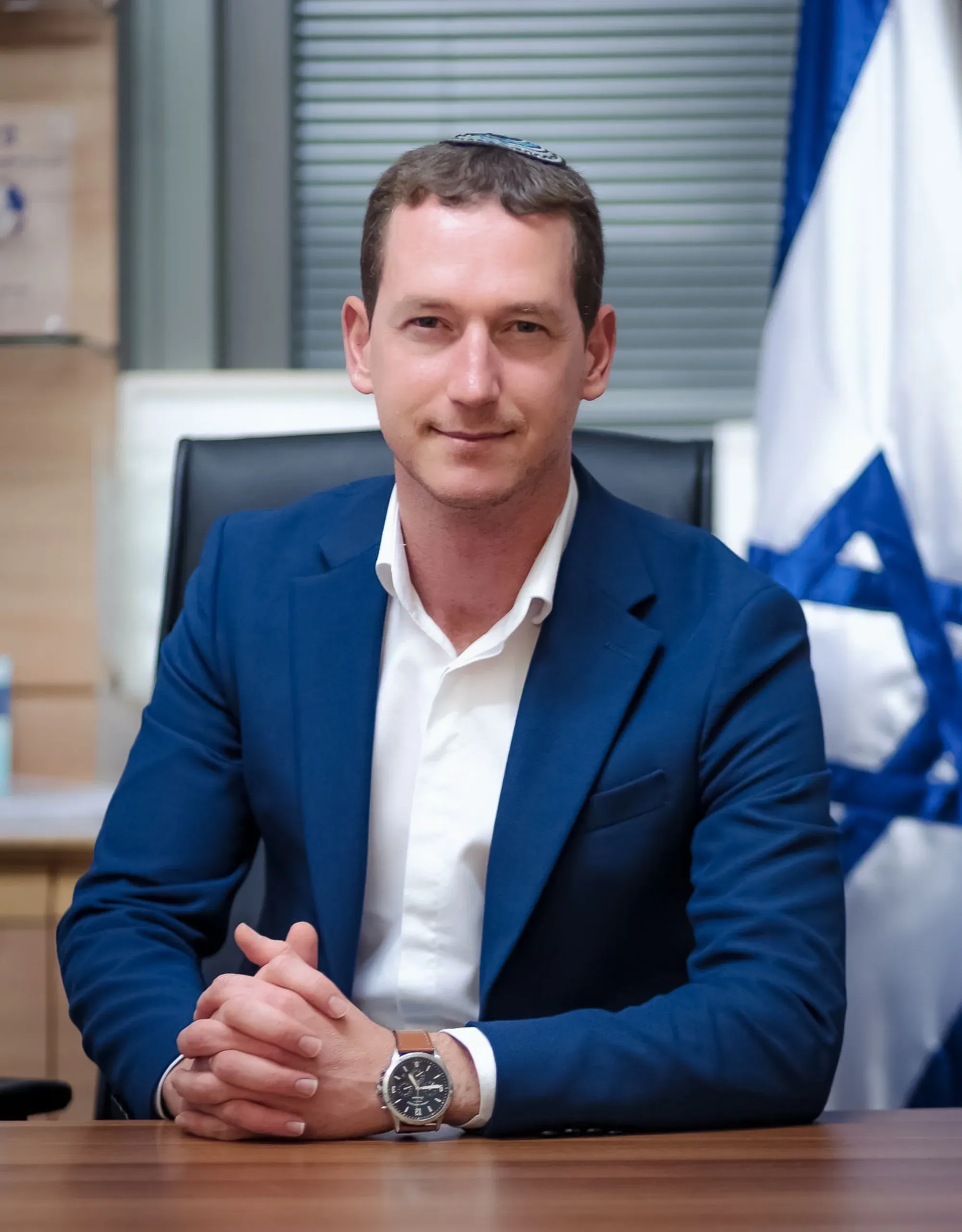 פורטרט עסקי של גבר בחליפה כחולה וכיפה, יושב ליד שולחן עם דגל ישראל