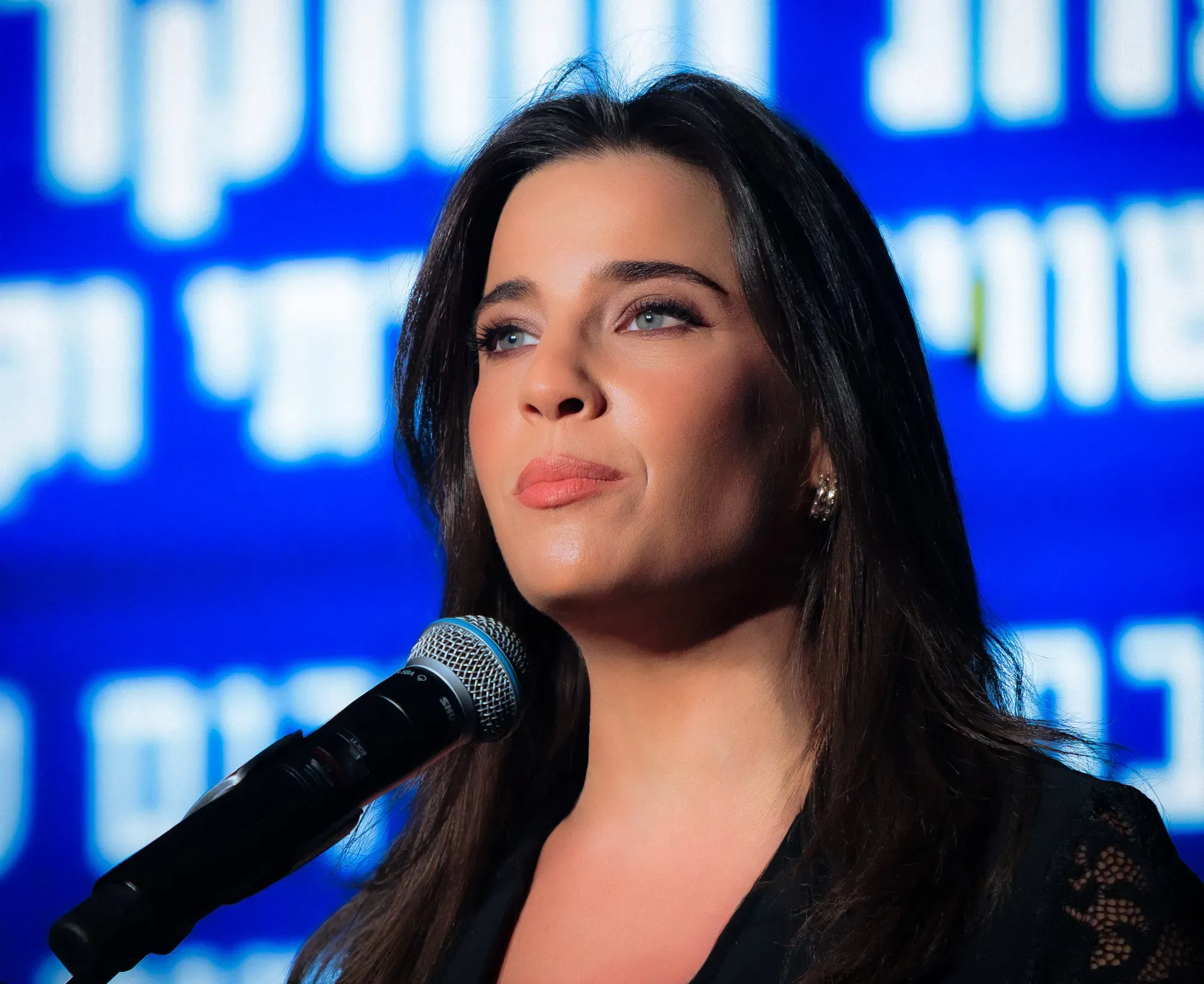 צילום אירוע: דניאל רוט-אבנרי מדברת מול מיקרופון על רקע מסך כחול
