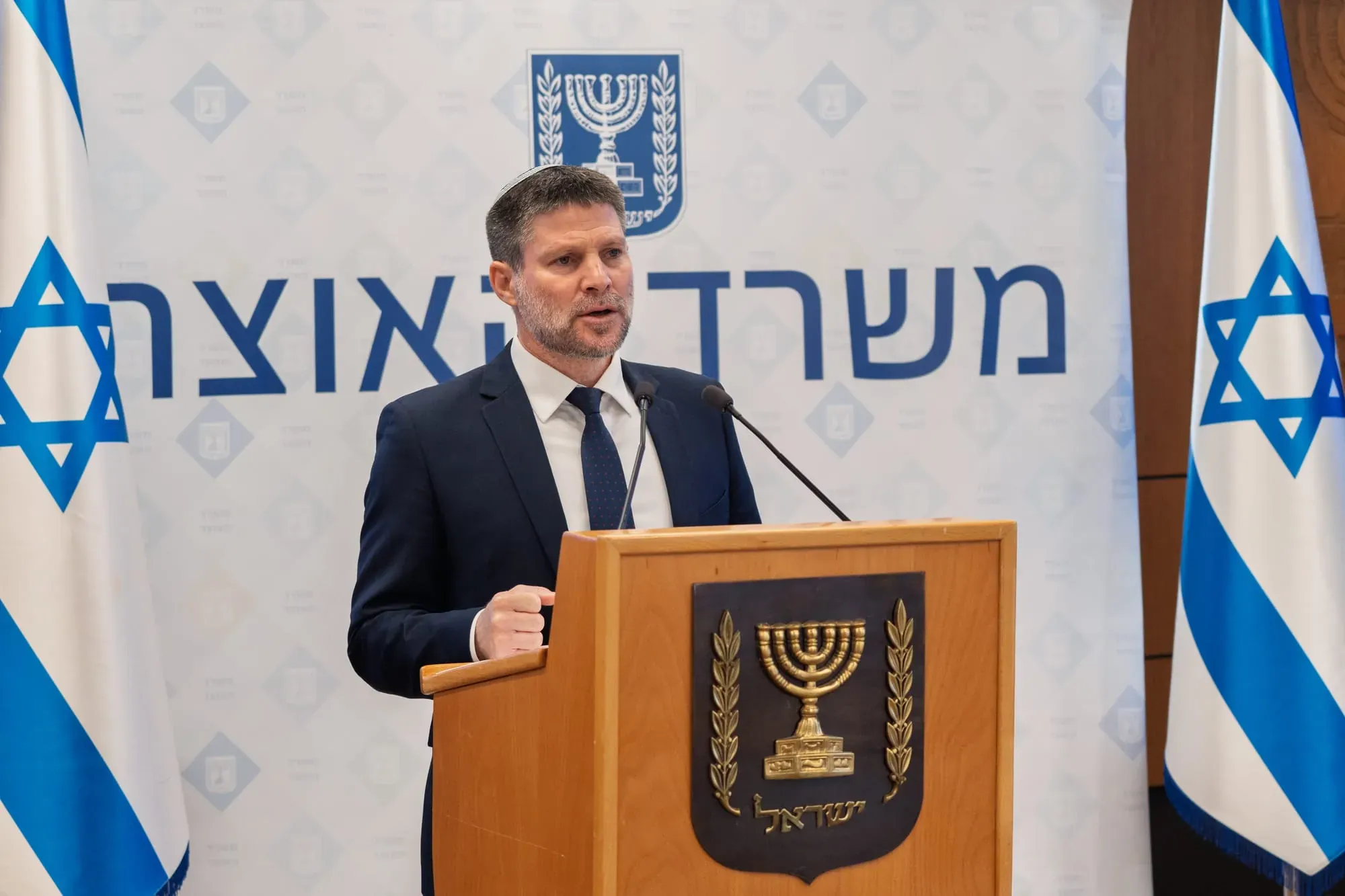 צילום אירוע: בצלאל סמוטריץ' נואם ליד דוכן עם סמל המדינה ודגלי ישראל