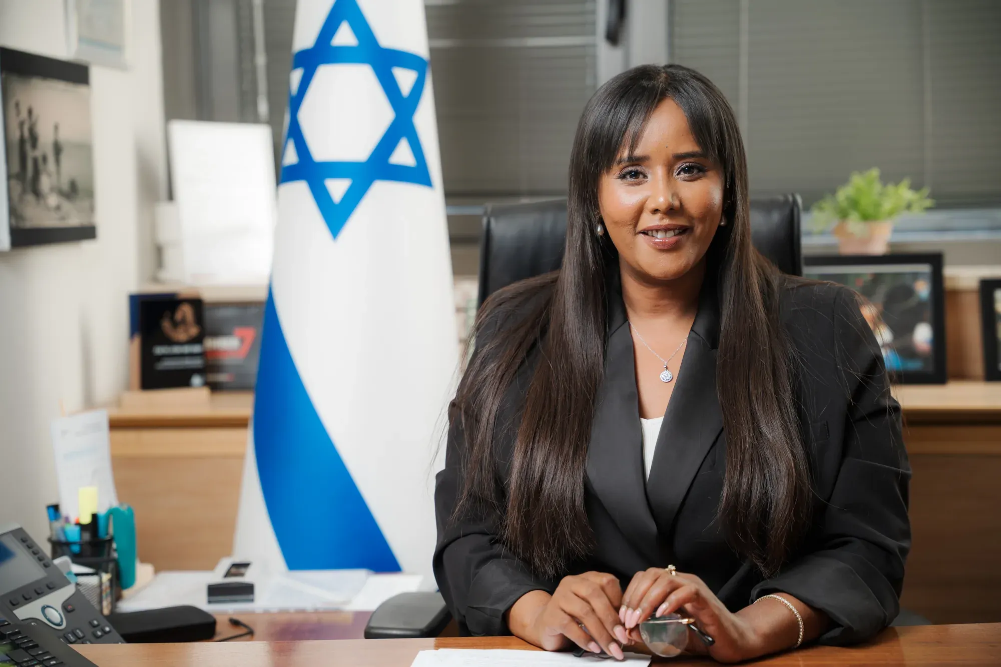 פורטרט עסקי של אשת ציבור בחליפה שחורה ליד שולחן עבודה ודגל ישראל