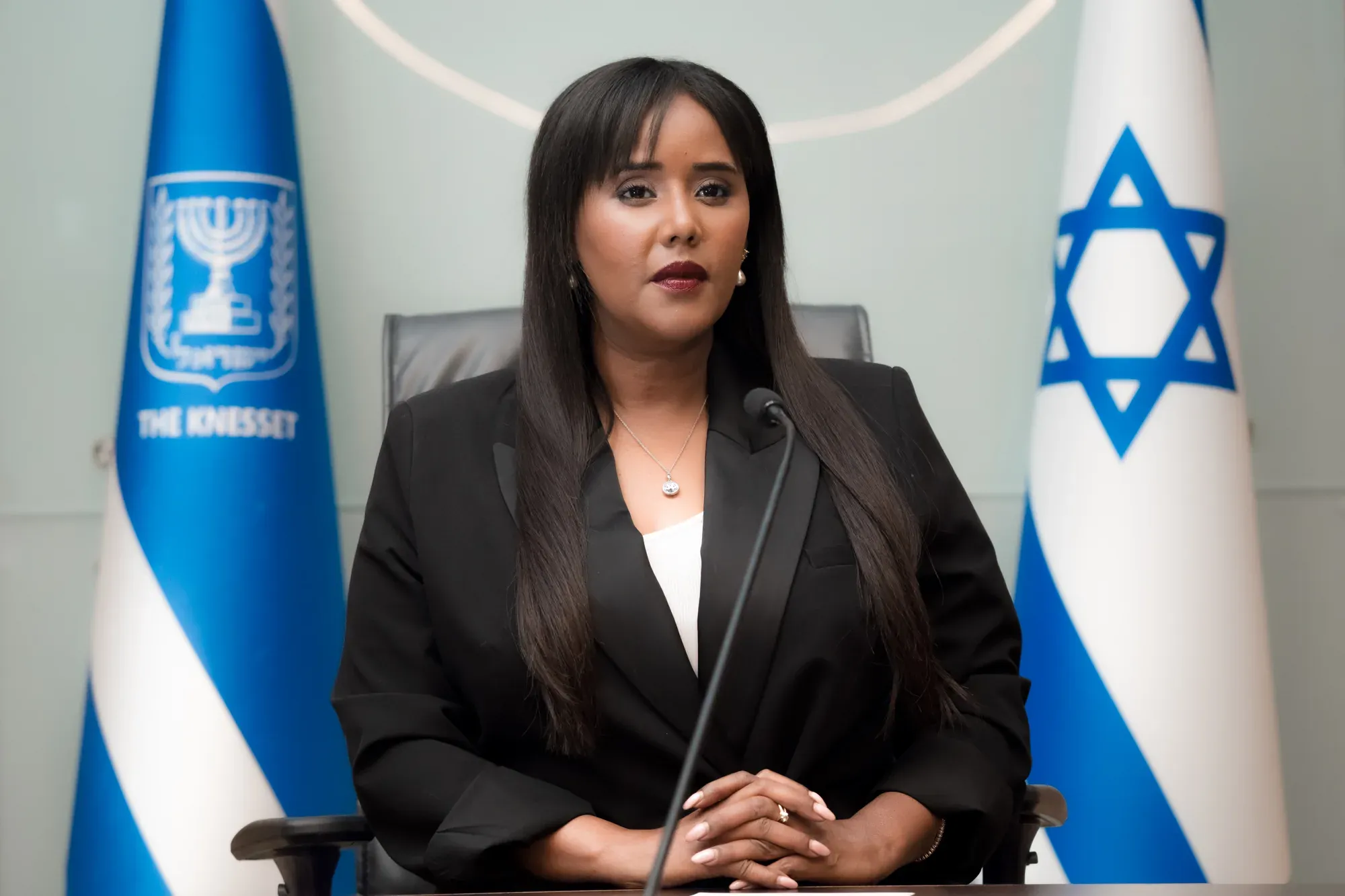 פורטרט רשמי של אשה בחליפה שחורה מאחורי מיקרופון, לצד דגל ישראל ודגל הכנסת