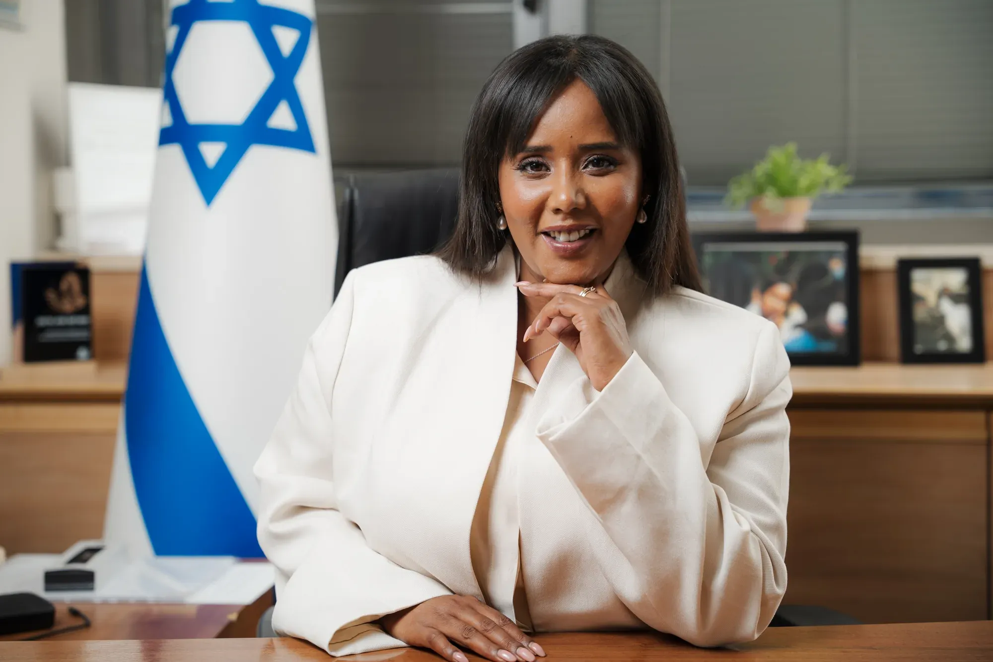 פורטרט של אשה בז'קט לבן ליד שולחן עבודה, יד אל הסנטר, ודגל ישראל מאחוריה